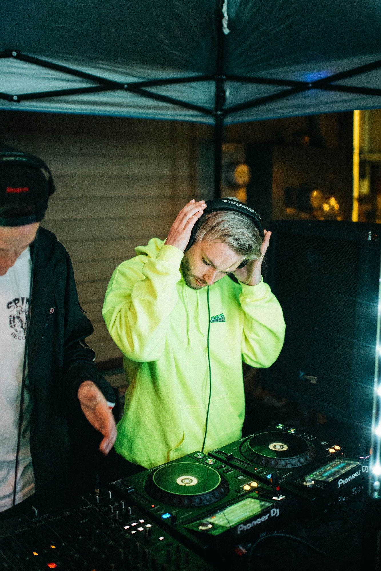 JohnLuc wearing a bright green sweatshirt and adjusting his headphones behind DJ equipment.